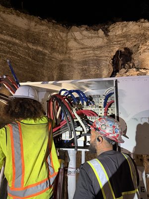 Two electricians working at night on an open electrical panel packed with blue, red and black conductors
