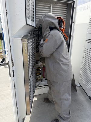 Technician in a full 55-cal arc-flash suit and hood working inside an open battery and inverter cabinet