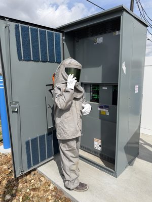 Technician in arc-flash PPE working at an open panel behind a safety barrier