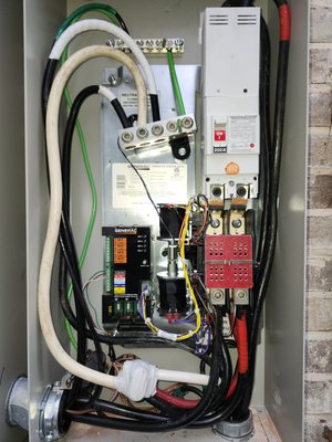 Open automatic transfer switch on a brick wall showing the 200-amp main breaker, neutral and ground bar, controller board and dressed conductors
