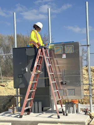 Crew member on a ladder atop a utility switchgear enclosure against a blue sky