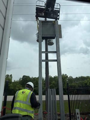 All American Electric worker in hi-vis up a ladder at a riser structure under a heavy storm sky