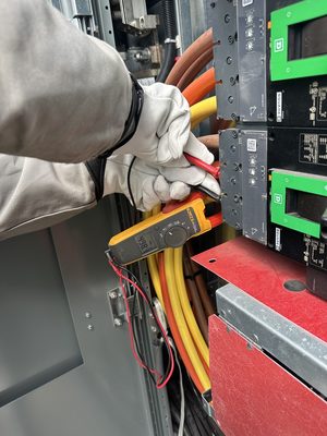 Gloved hand holding an electrical tester against the conductors landed on a switchgear breaker, behind a red arc-flash barrier