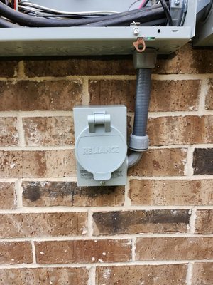 Weatherproof generator power inlet box mounted on a brick wall with flexible conduit running up to the panel