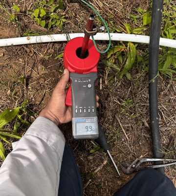 Electrician holding a clamp-on ground resistance tester around a driven ground rod, the display reading 9.9 ohms, with the test set open on the ground beside it