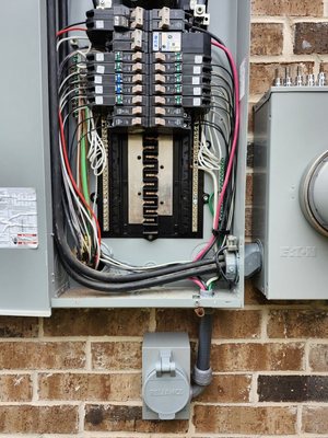 Open residential load center on a brick wall with breakers landed and conductors dressed, the meter can beside it and a generator power inlet mounted below