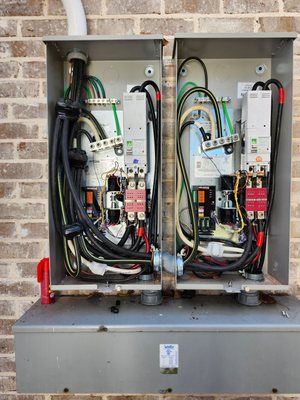 Two automatic transfer switch enclosures mounted side by side on a brick wall, both open, each with its own controller and contactor