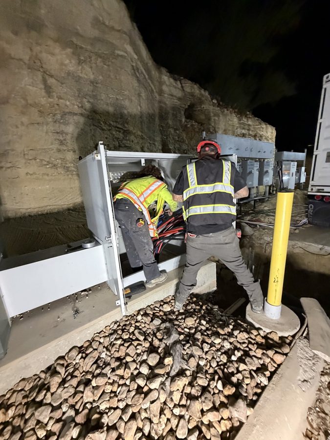 Two All American Electric electricians in hi-vis vests working inside an open meter and disconnect cabinet at night