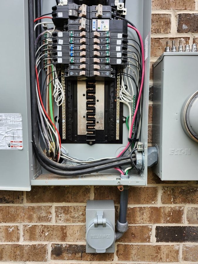 Open residential load center on a brick wall with breakers landed and conductors dressed, the meter can beside it and a generator power inlet mounted below