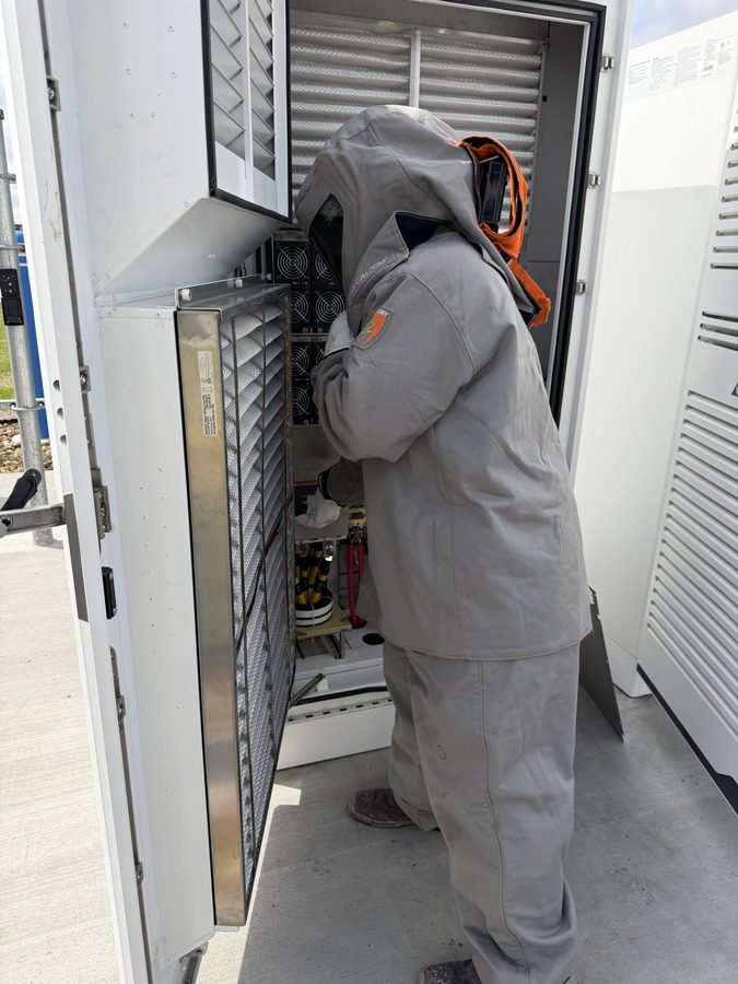 Technician in a full 55-cal arc-flash suit and hood working inside an open battery and inverter cabinet